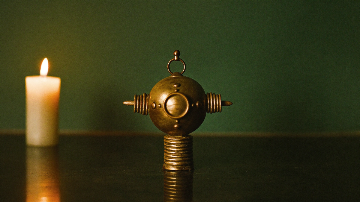 Small brass sphere on a coiled stand beside a lit candle against a dark green wall