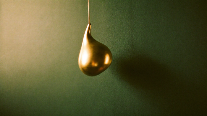 A polished brass plumb weight hanging motionless from a fine cord against a dark green wall