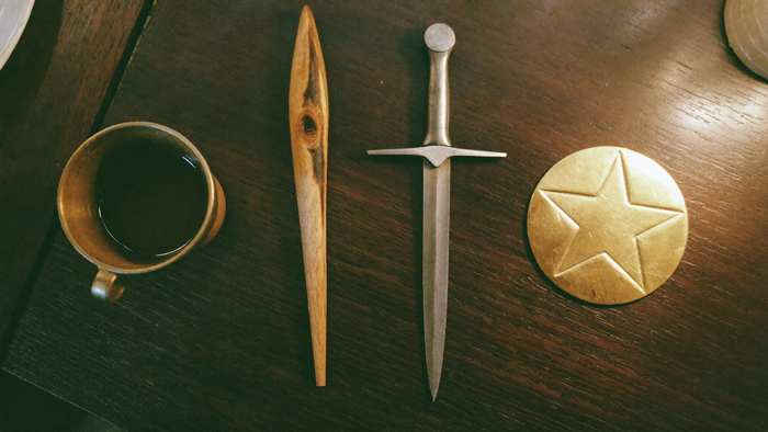 The four tarot suits as objects on dark walnut: a pewter cup, a wooden wand, a steel dagger and a gold pentacle disc