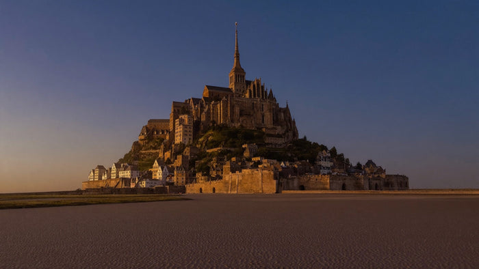 Mont-Saint-Michel at dusk: granite tidal island with Gothic abbey and spire above rippled sand flats at low tide