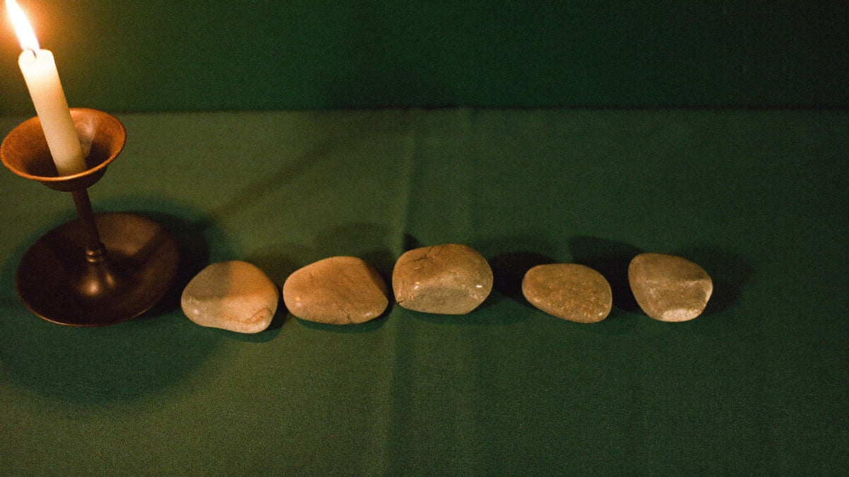 Five smooth pale stones laid in a row on dark green linen beside a single burning candle in a plain holder
