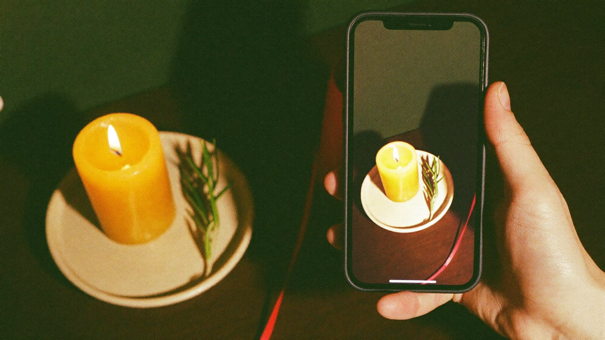 Hand holding a smartphone that photographs a burning beeswax candle with rosemary and red thread on a small altar
