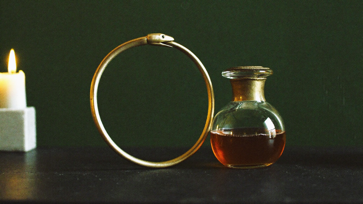 A bronze snake biting its own tail forming a closed ring, beside a round glass bottle of amber liquid and a white candle