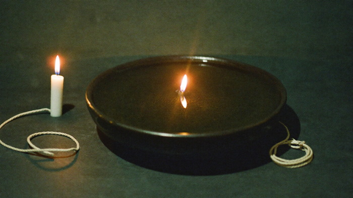 A dark bowl of still water reflecting a single candle flame beside a coil of raw linen cord