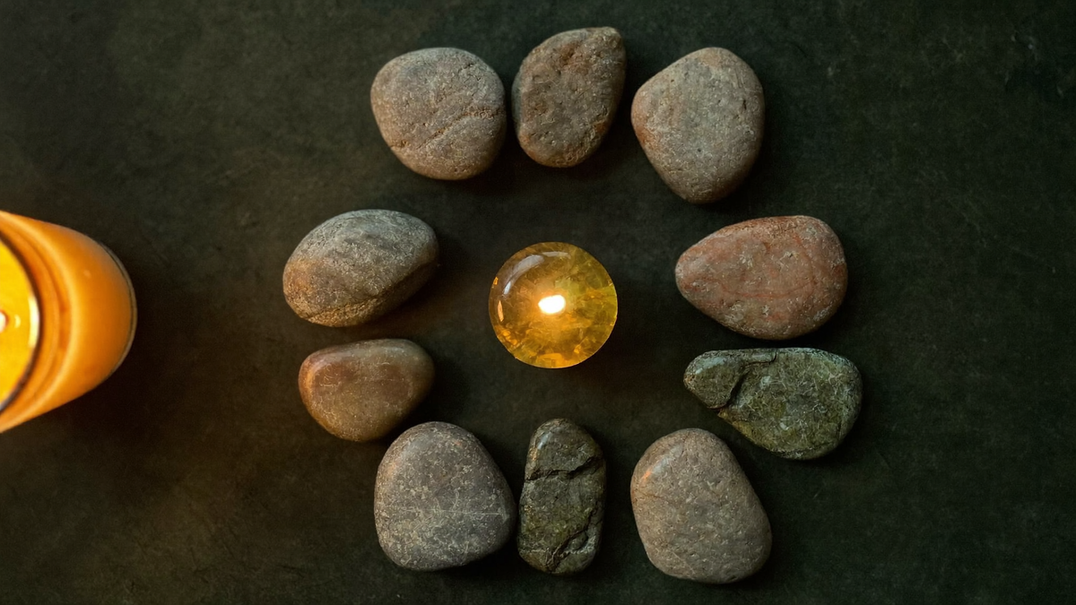 Ring of smooth river stones on dark slate around a single glowing amber stone, warm candlelight from the left edge