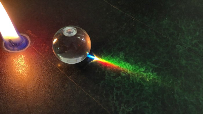 A clear glass sphere on dark stone splitting candlelight into a band of spectrum colours