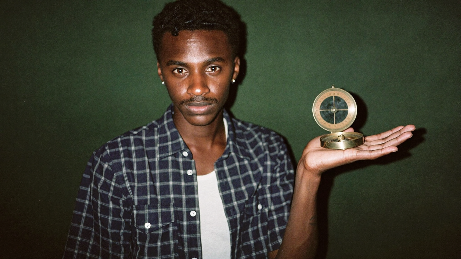 Young man holding a brass compass flat in his open palm against a dark green wall
