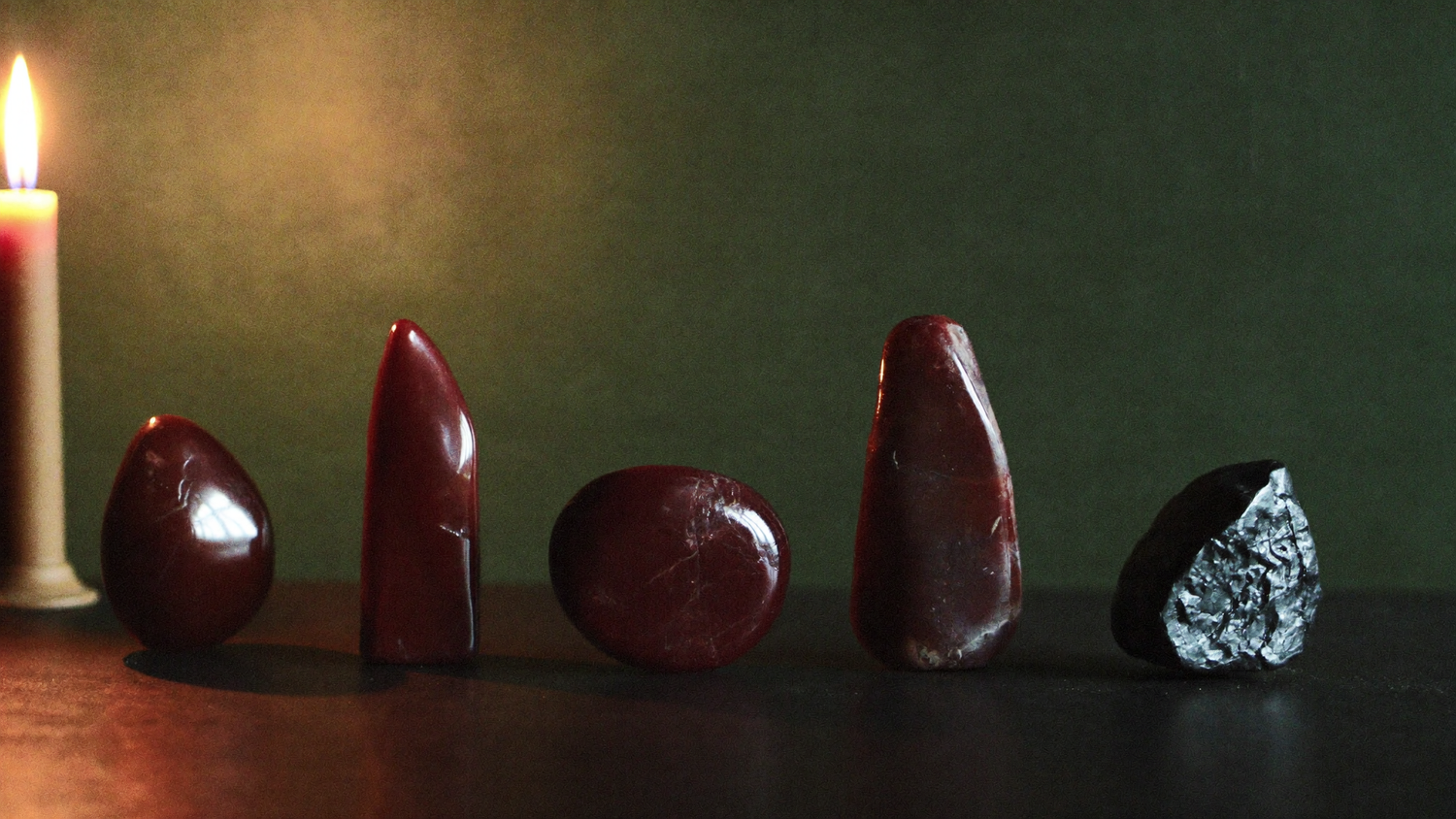 Four polished deep red stones and one dark crystal in a row on slate beside a lit candle