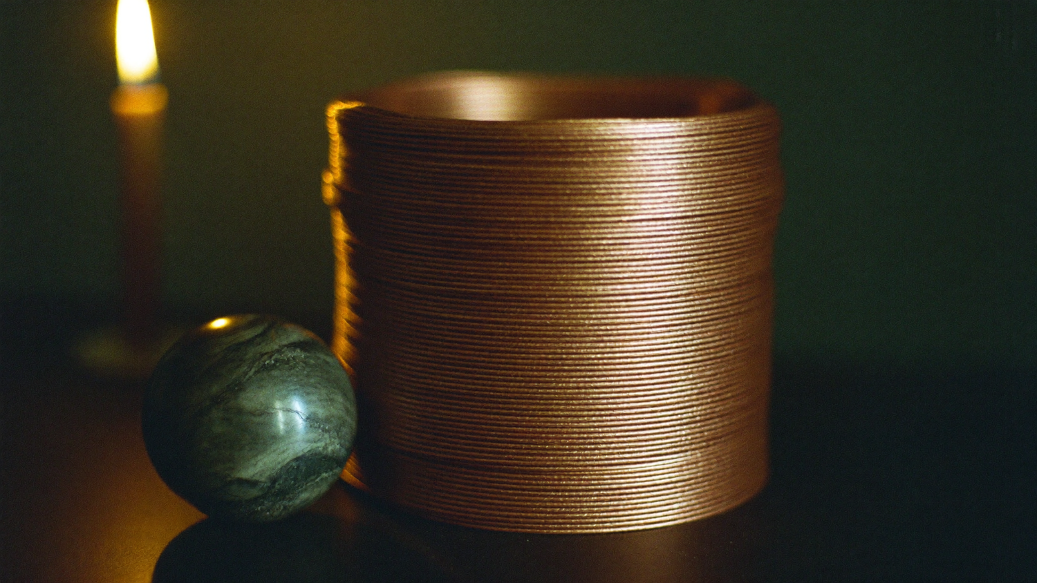 Coiled copper wire beside a marbled stone sphere on dark stone, lit by a single candle