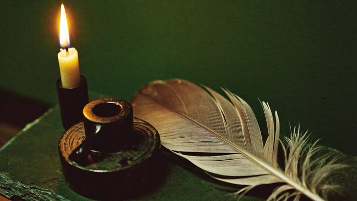 Grey goose feather quill and a dark horn inkwell beside a burning candle on a dark green stone slab
