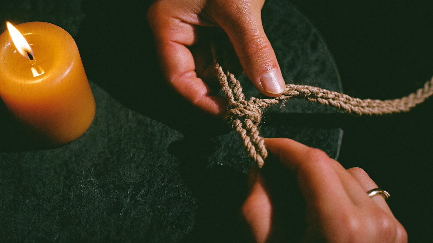 Two hands untying a knot in a length of raw linen cord by candlelight on a dark stone surface.