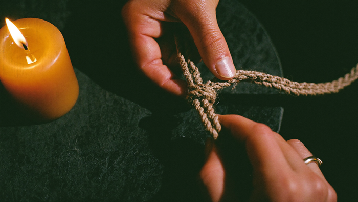 Two hands untying a knot in a length of raw linen cord by candlelight on a dark stone surface.