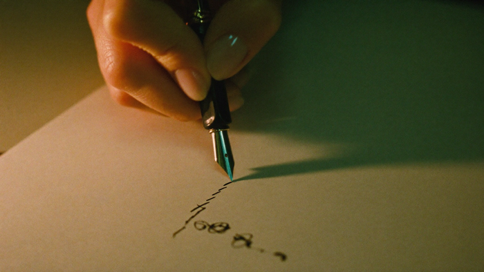 A hand writing in dark ink on cream paper with a fountain pen under warm candlelight