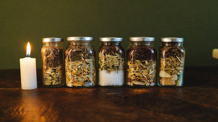 Five sealed spell jars filled with layered dried herbs, salt and stones on dark wood beside a lit candle