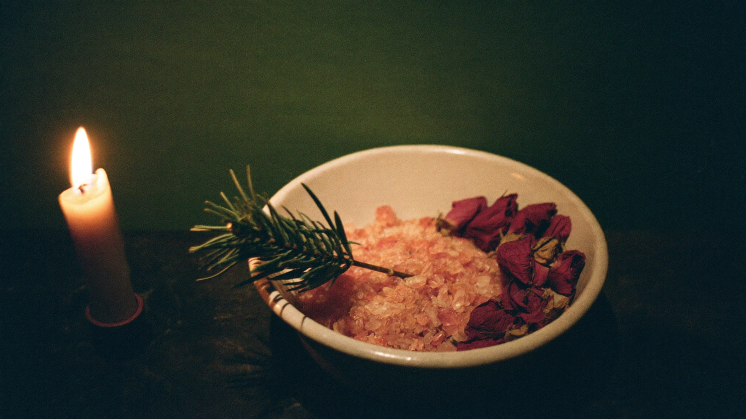 Bath salt with dried rose petals and a fir sprig in a ceramic bowl beside a burning candle