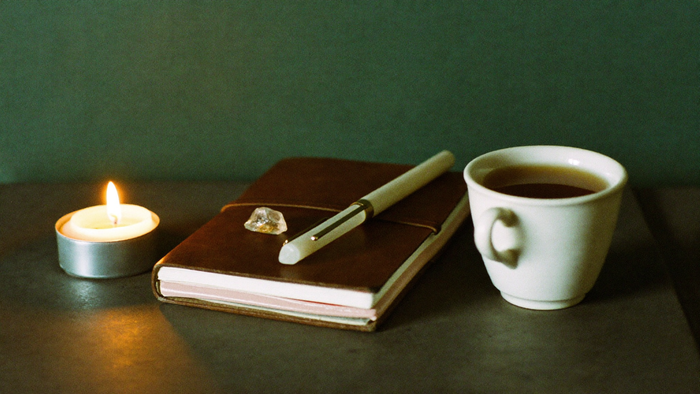 Open journal with a pen and clear quartz beside a lit tealight on a dark green desk
