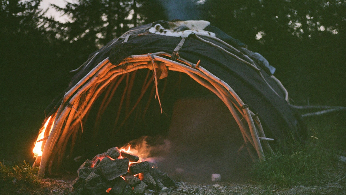 Willow-frame sweat lodge dome draped in dark blankets at dusk, with a pile of glowing stones and fire at the entrance