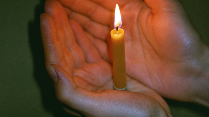 Two cupped hands sheltering the flame of a burning beeswax candle in the dark