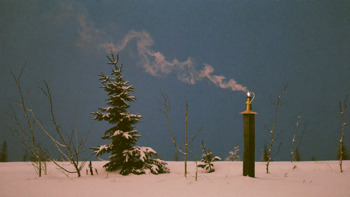 Snow-covered winter field at blue dusk with a lantern flame and drifting smoke on a wooden post
