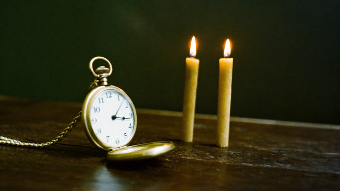 Open brass pocket watch on dark wood beside two lit beeswax candles