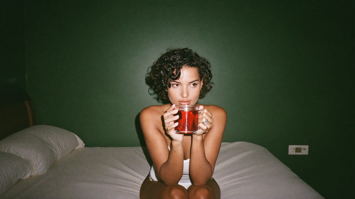 Young woman sitting alone on the edge of a bed holding a mug against a dark green wall