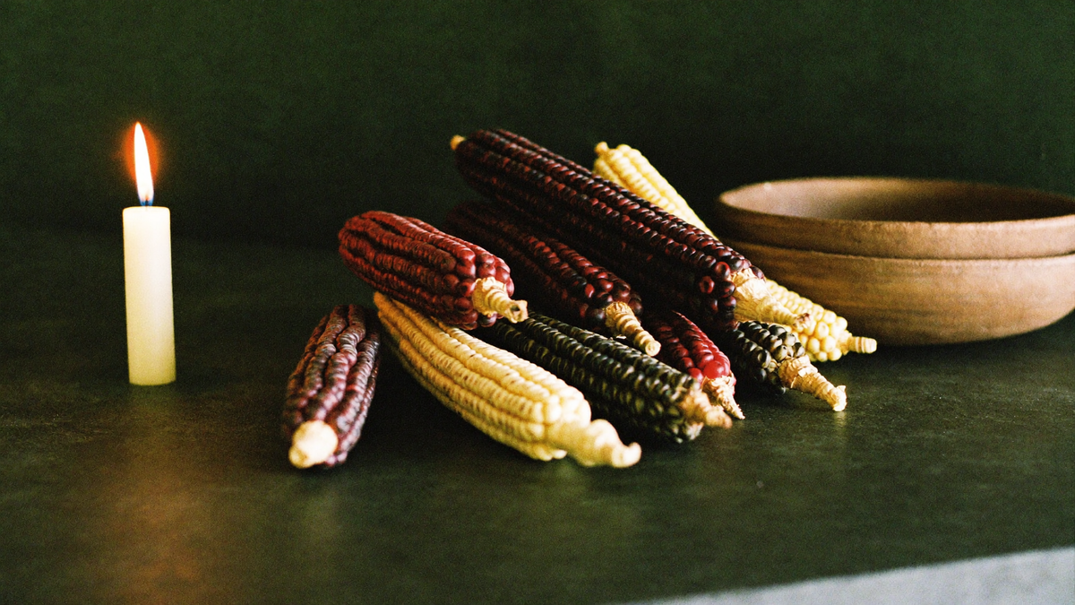 Dried ears of red, black and pale yellow maize beside a clay bowl, lit by a single candle