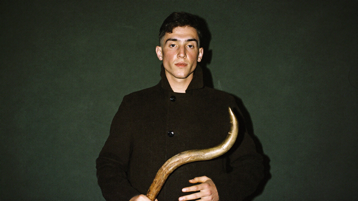 A young man in a dark wool coat holds a large curved horn in front of a deep green wall