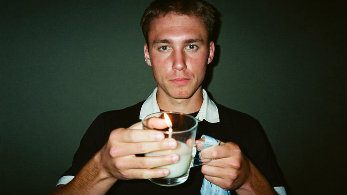 A young man holding a lit white candle in a glass against a dark green wall