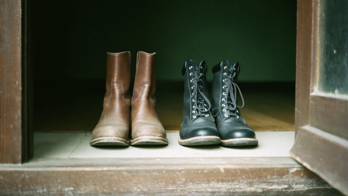 Two pairs of boots side by side on a wooden threshold, one worn brown leather pair and one dark laced pair, soft light