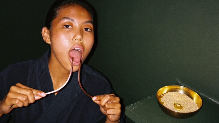 Woman in a dark robe using a copper tongue scraper beside a brass bowl of sesame seeds and oil