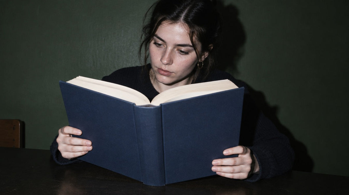 Young woman reading a thick open book with a plain dark blue cover, studying at a table by a green wall