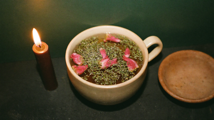 Cup of herbal infusion scattered with rose petals beside a lit candle and a shallow clay dish on a dark table