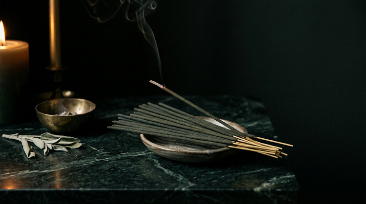 Hand-rolled incense sticks in a ceramic ash catcher on dark green marble, one lit with rising smoke, beside a brass bowl and dried sage in candlelight