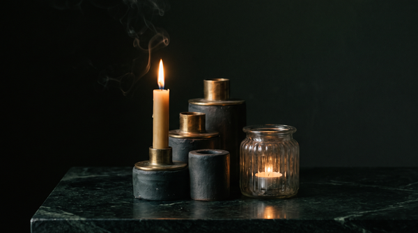 A stepped group of handmade ceramic and marble taper candle holders beside a fluted glass lantern on dark green marble, one holding a lit beeswax taper, lit by warm candlelight