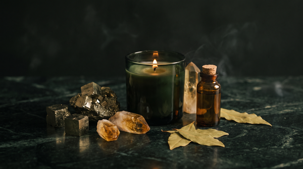 Raw golden pyrite cubes, honey-colored citrine points, a dark green glass ritual candle, an amber spell-oil bottle and dried bay leaves on dark green marble, lit by candlelight