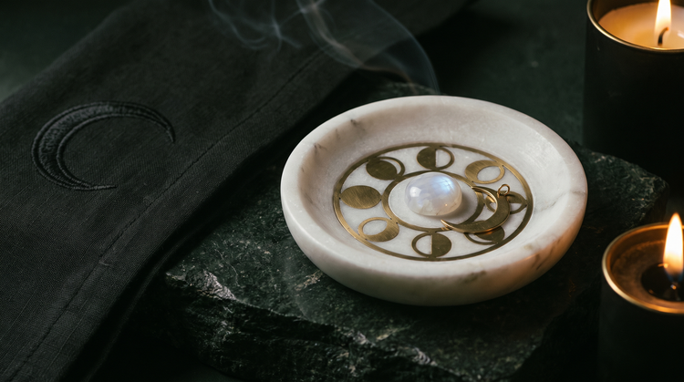 A hand-carved white marble incense dish with brass moon-phase inlay, a pale moonstone cabochon and a brass crescent charm on dark stone beside a black crescent-embroidered cloth, lit by candlelight