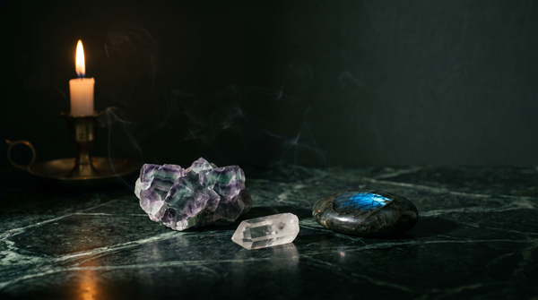 Banded purple and green fluorite beside a clear quartz point and a dark labradorite palm stone flashing blue, on dark green marble lit by a single candle