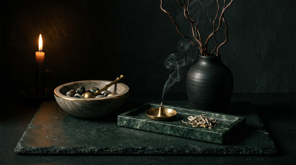 A carved stone bowl of tumbled stones, a dark green marble tray with a brass incense holder and loose sage, and a matte black ceramic vase with bare branches, on dark slate lit by a single candle