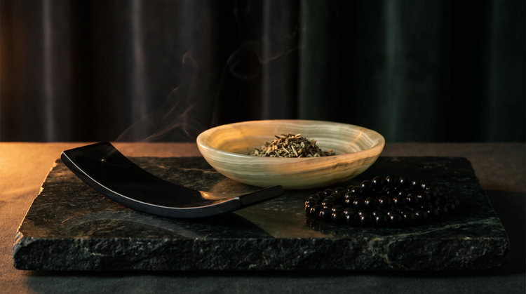 A carved black onyx incense rest, a banded cream onyx bowl glowing at the rim and a strand of black onyx beads on dark green marble in candlelight