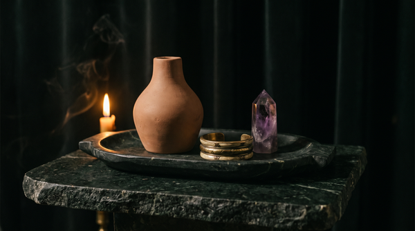 A dark marble tray with a terracotta vase, a brass cuff bracelet and a polished amethyst point on dark green stone in candlelight