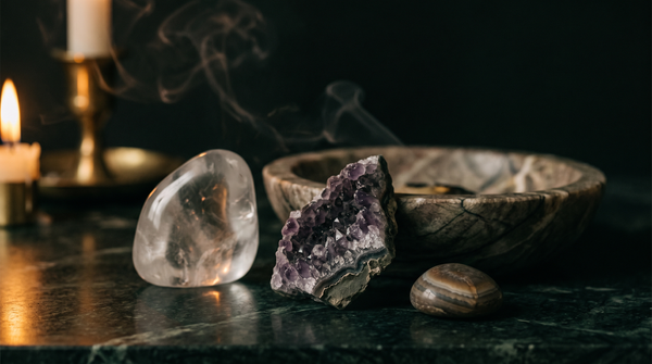 A polished clear quartz freeform, a raw amethyst geode fragment and a banded tumbled pebble beside a veined stone bowl on dark green marble, lit by a single candle with drifting smoke