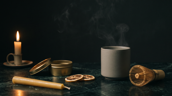 A steaming unmarked stoneware cup, a bamboo whisk, an open brass tin, dried citrus slices and an unlit beeswax taper on dark green-black marble, lit by a single candle from the left