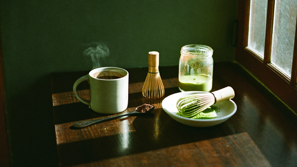 A steaming mug of cacao beside bamboo whisks and a bowl of matcha on a dark wood counter in morning light
