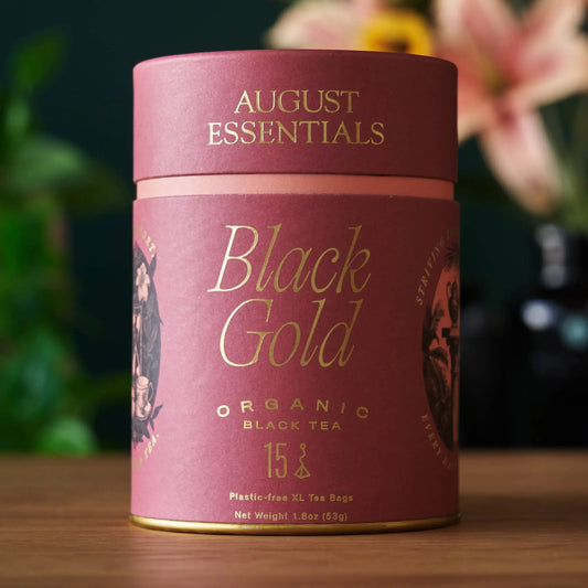 A pink August Essentials carton of Black Gold organic black tea, 15 tea bags, on a wooden surface.
