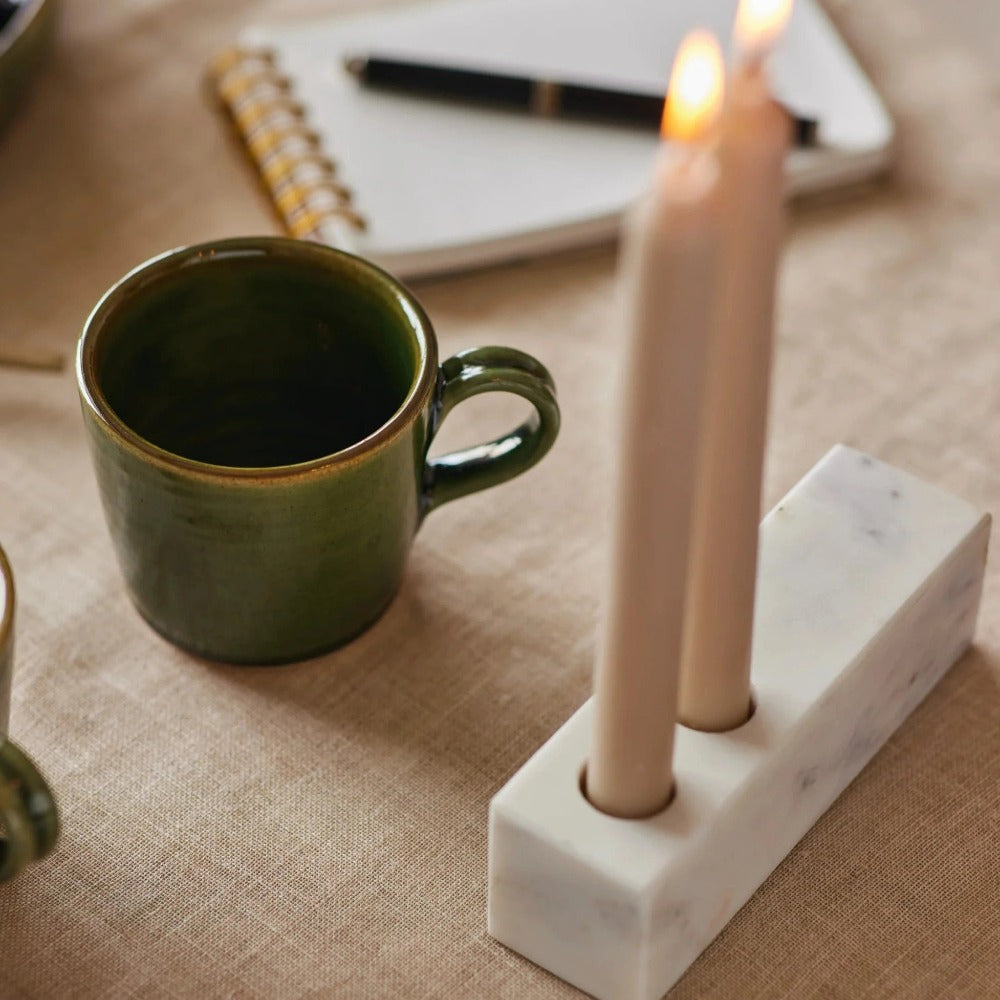 Marble candle holder with lit tapers beside a green mug and a notebook