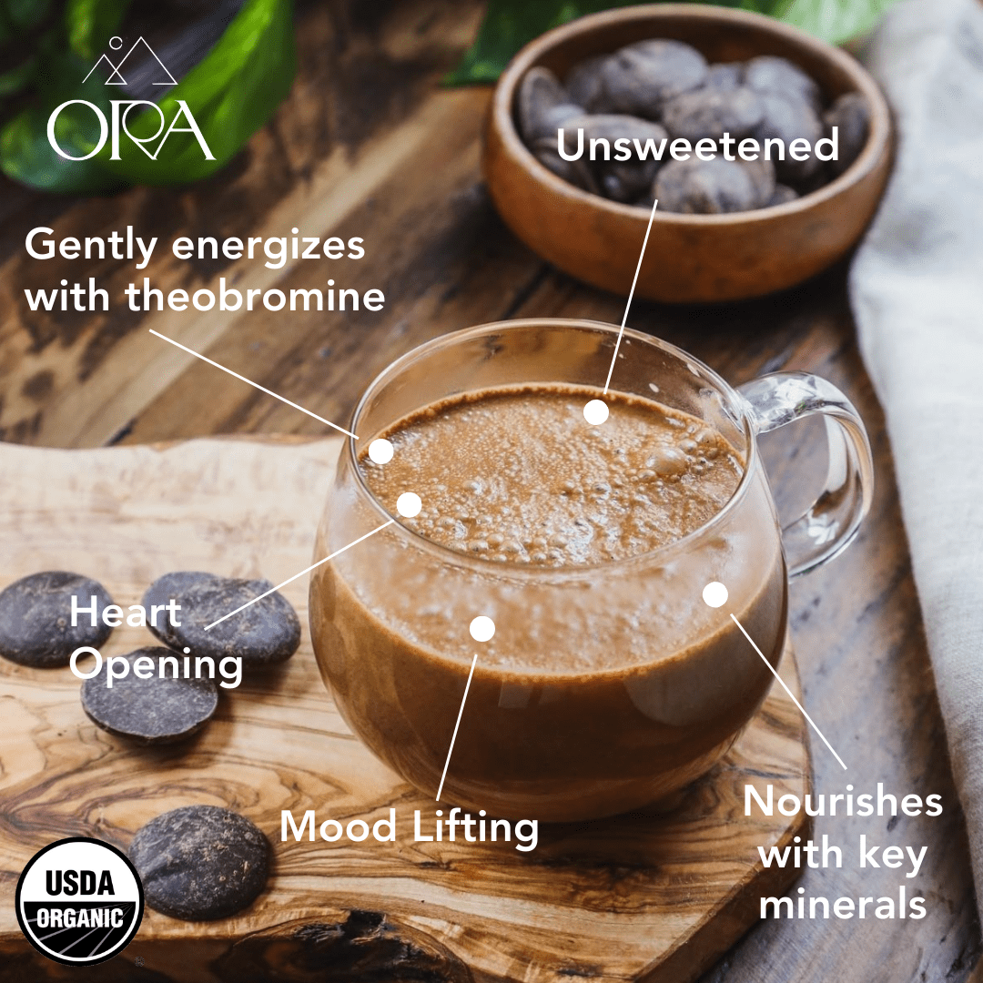 Glass mug of whisked Ora ceremonial cacao on a wooden board with cacao discs, labelled unsweetened, gently energizing and nourishing