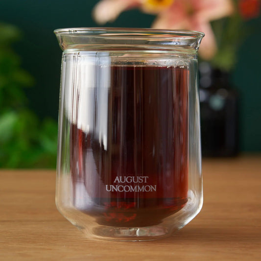A double-walled hand-blown glass holding brewed amber tea, marked August Uncommon, on a wooden surface.