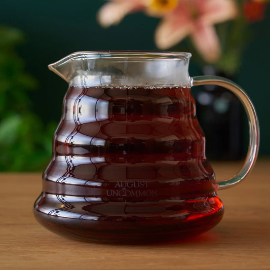 A heatproof glass August Uncommon teapot filled with brewed amber tea, on a wooden surface.