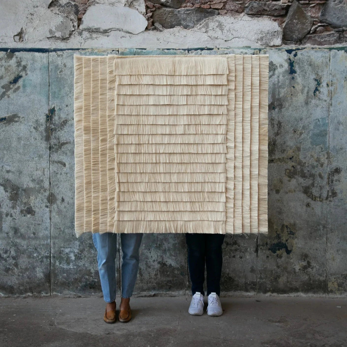 Two people holding the square Campo de Arroz tapestry against a weathered wall
