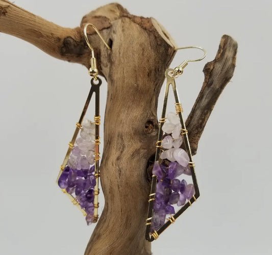 A pair of raw amethyst drop earrings in gold-tone wire settings, hanging from a piece of driftwood.